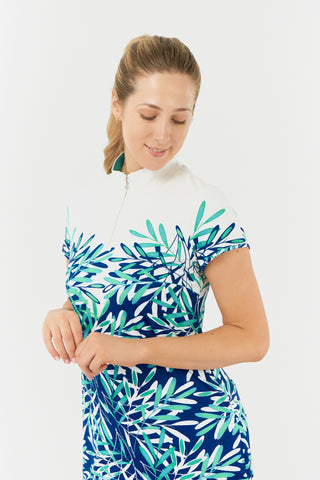 The close-up model shot of the Pure Golf Carly Cap Sleeve dress features an aquatic design in a blend of blue and ocean tones, highlighted by a floating palm print from the chest down. This longer-length dress is crafted from 4-way stretch fabric for maximum flexibility and comes with detachable shorts for added coverage. Its flattering fit suits all body types, while the mock neck collar adds a sleek, modern flair. Designed to resist creasing, static, and moisture, the dress also offers UPF 50+ protection.
